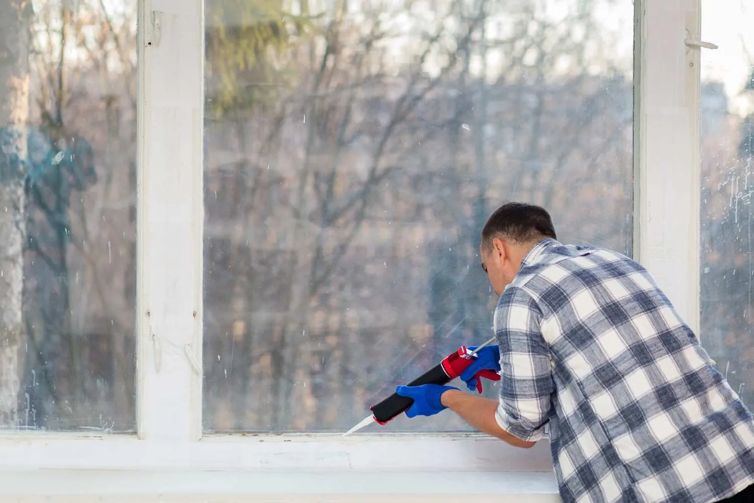 Man in a plaid shirt sealing a white window frame with a caulk gun